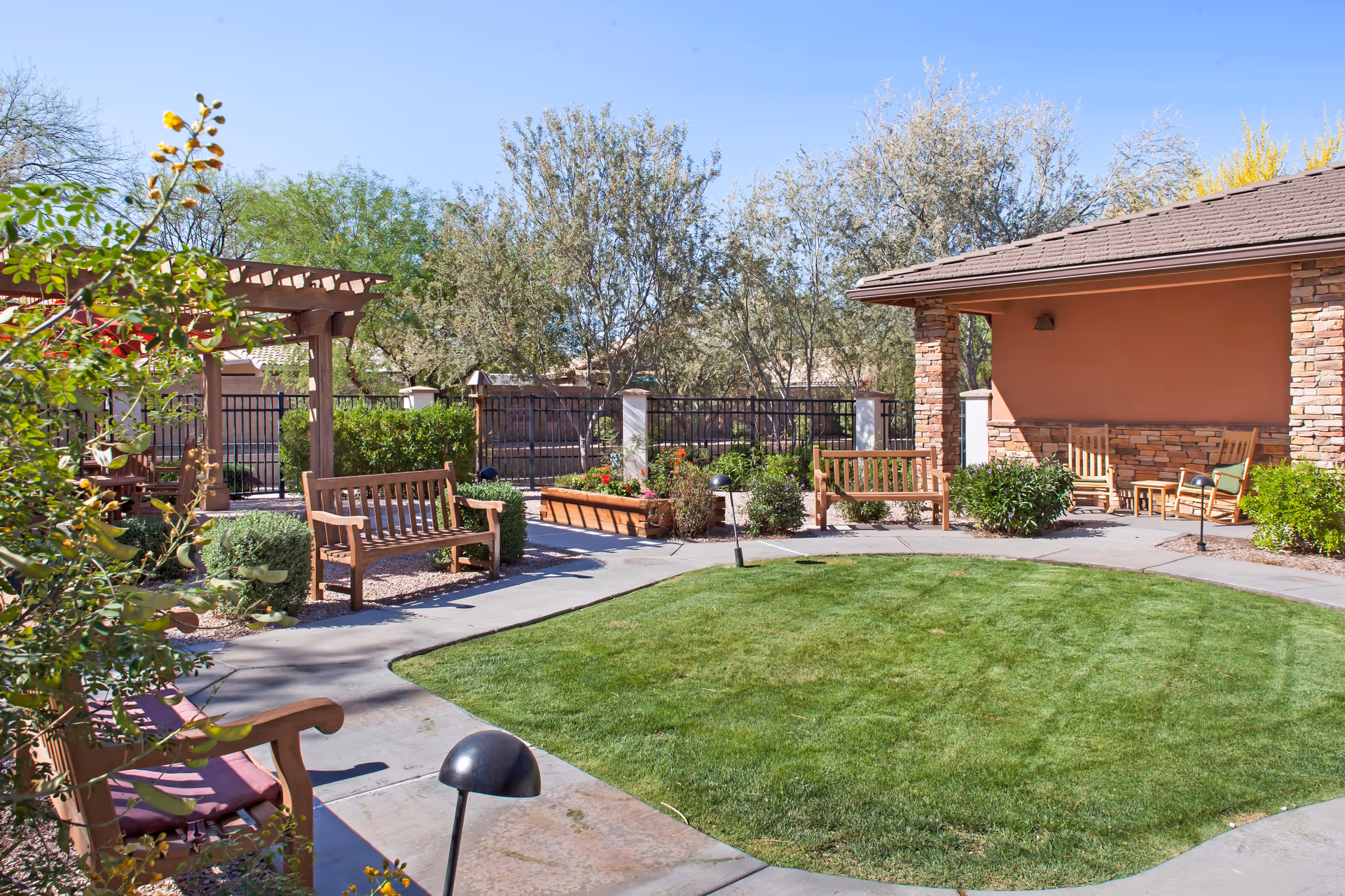 Outdoor garden area at Sunrise of Chandler featuring a circular patch of green grass surrounded by a concrete walkway. Wooden benches and chairs are placed around the garden, with some under a pergola and others near a covered patio with stone pillars. Various shrubs, plants, and trees are visible, along with a clear blue sky.