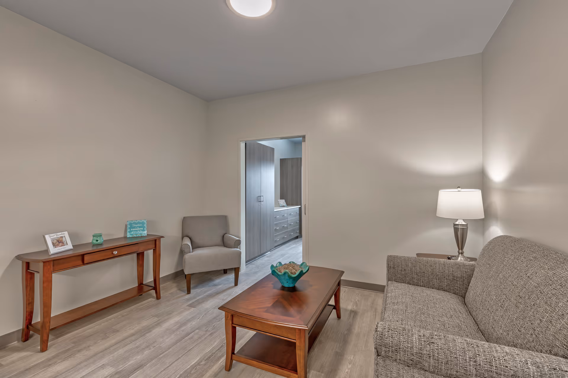 A small living room with a gray upholstered sofa and armchair, a wooden coffee table with a decorative bowl, a wooden console table with framed photos and a candle, and a table lamp on a side table. The room has light-colored walls and wood flooring, with an open doorway leading to another room with cabinets and drawers.