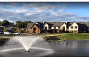 View of a senior living building across a pond with a fountain spraying water under a partly cloudy sky.
