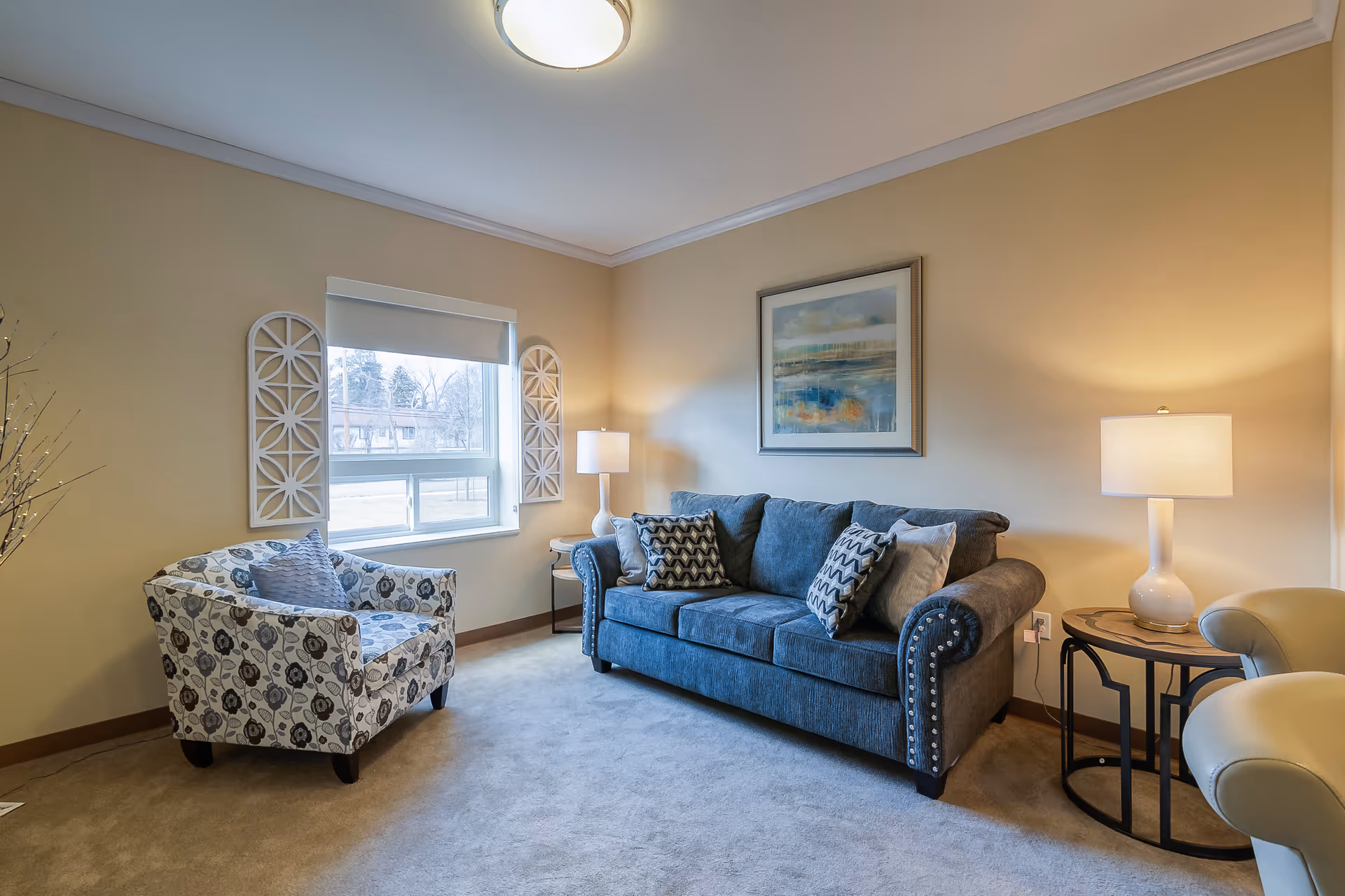 Cozy living room with a blue sofa, patterned armchair, side tables with lamps, wall art, and a window.