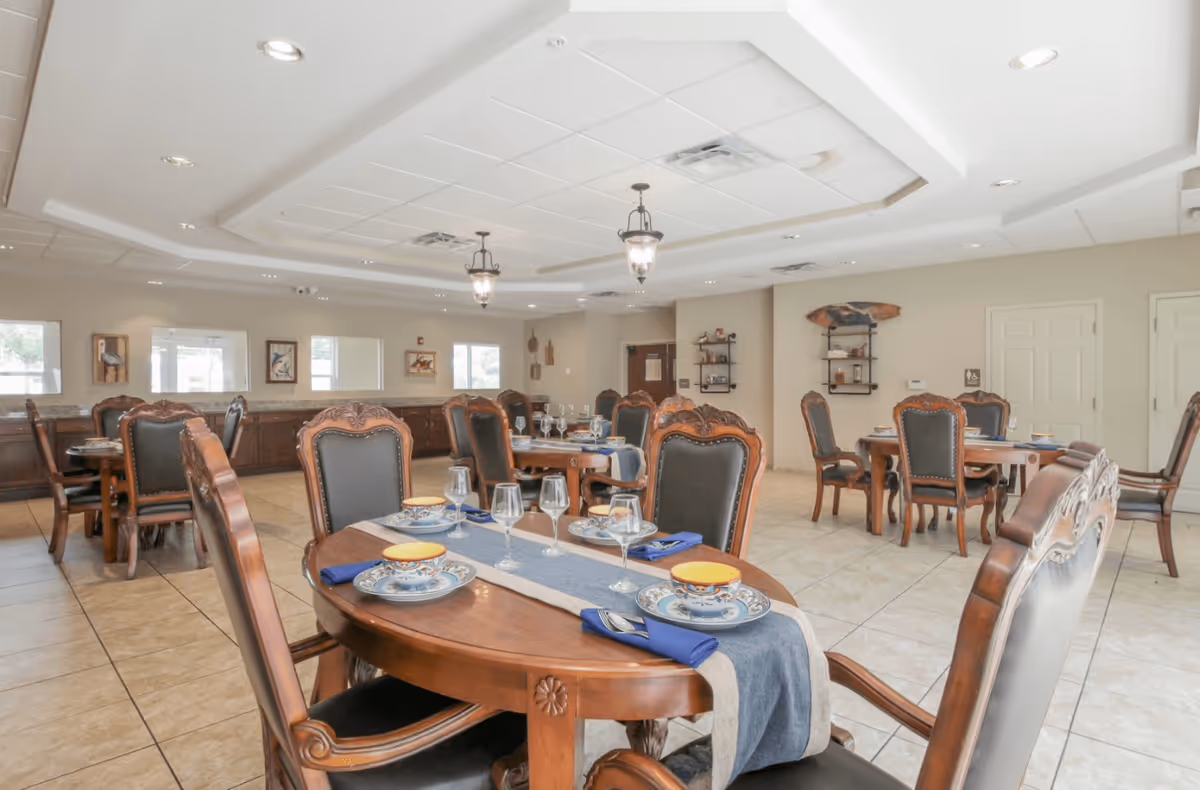 A spacious dining room with several wooden tables and chairs arranged neatly. Each table is set with plates, bowls, glasses, and blue napkins. The room has tiled floors, beige walls, decorative wall art, and ceiling lights with hanging lantern-style fixtures.
