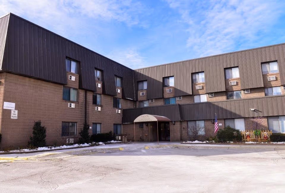 Brick senior living building with entrance canopy, American flag, and parking lot in foreground.