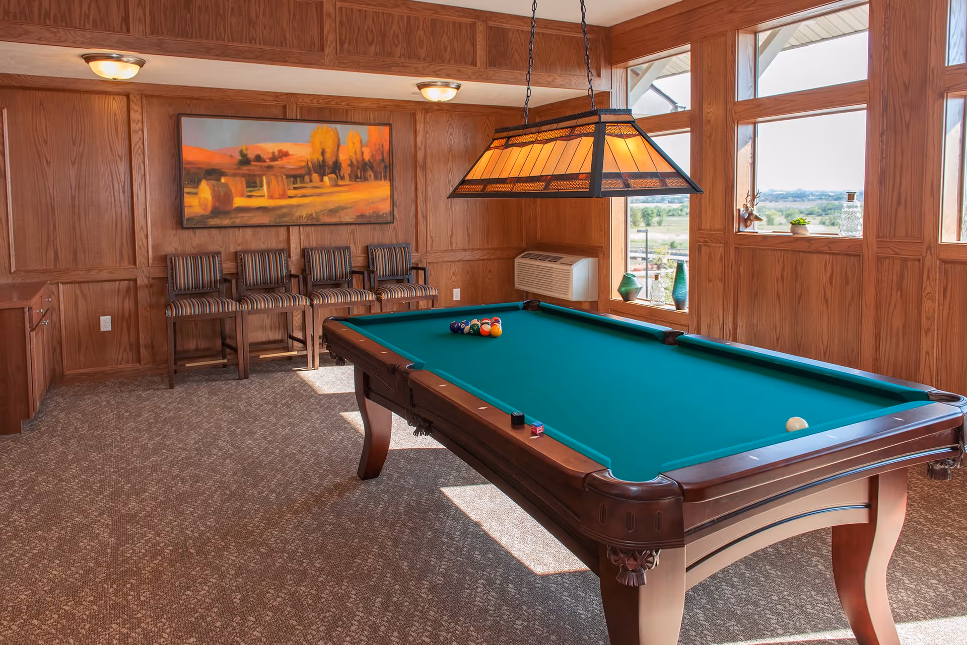 A well-lit game room with a green felt pool table in the center, a hanging stained glass light fixture above it, four striped chairs lined up against a wood-paneled wall, and large windows letting in natural light with a view outside. A landscape painting hangs on the wall above the chairs.