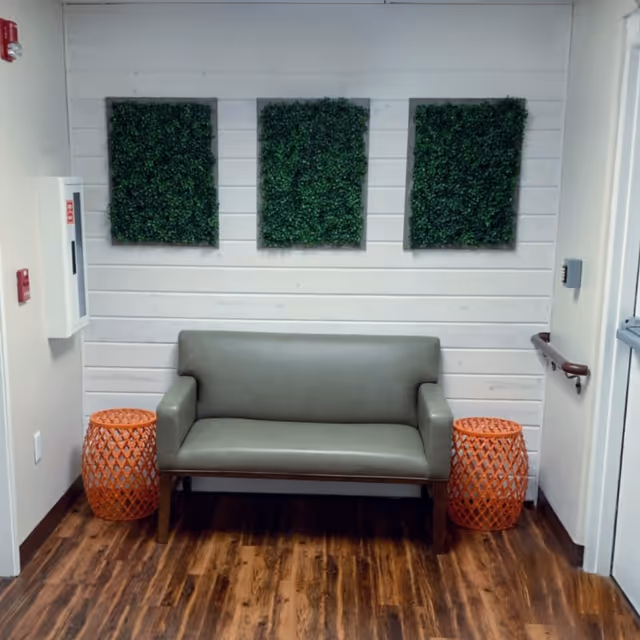 Small seating area with a gray loveseat between two orange side tables beneath three square green wall panels on a white shiplap wall.