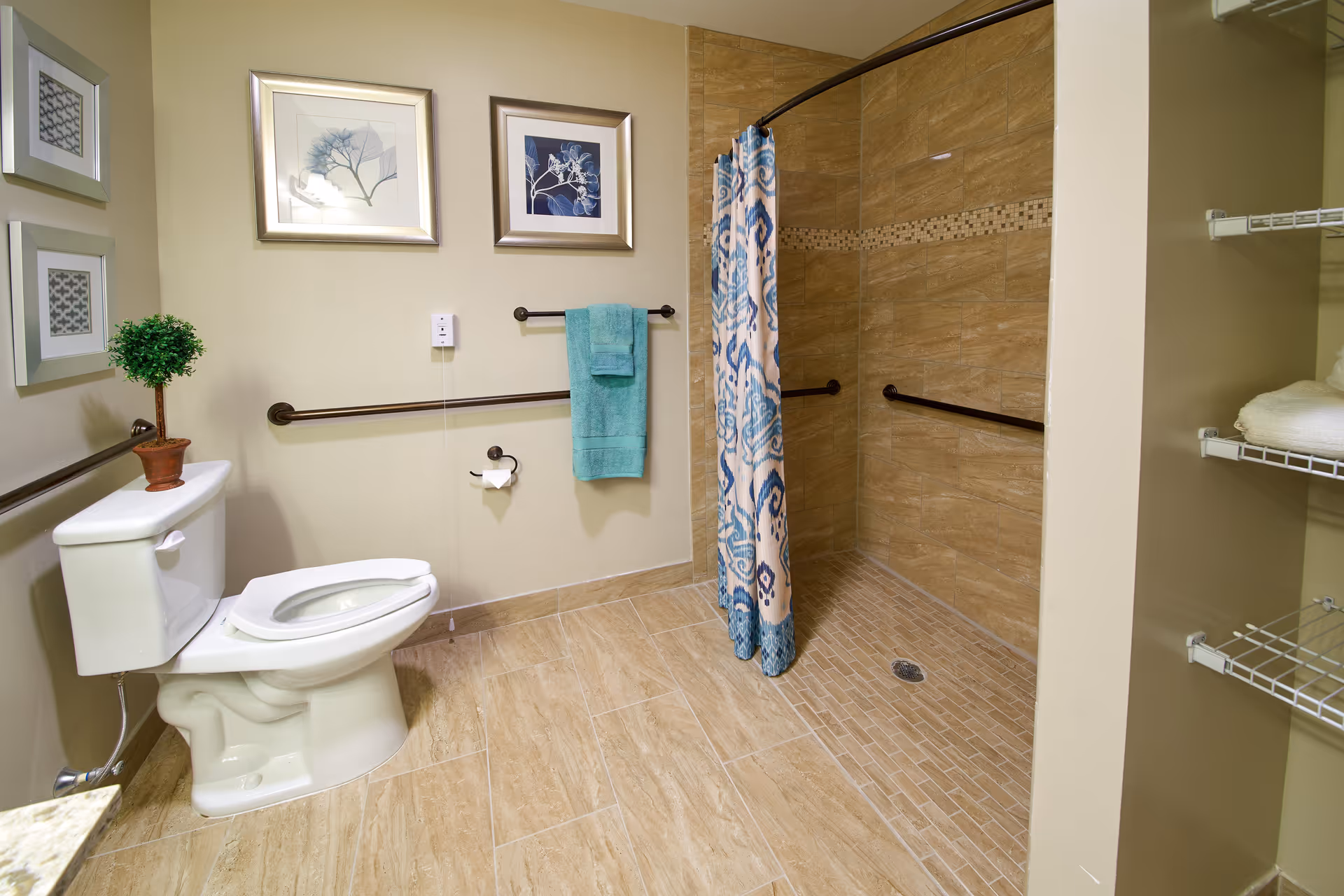 A spacious bathroom with beige tiled floor and walls. There is a white toilet with a small potted plant on the tank. The shower area has a patterned blue and white curtain and grab bars installed on the walls. Two framed pictures hang on the wall above a towel rack with a teal towel. Shelving with folded white towels is visible on the right side.
