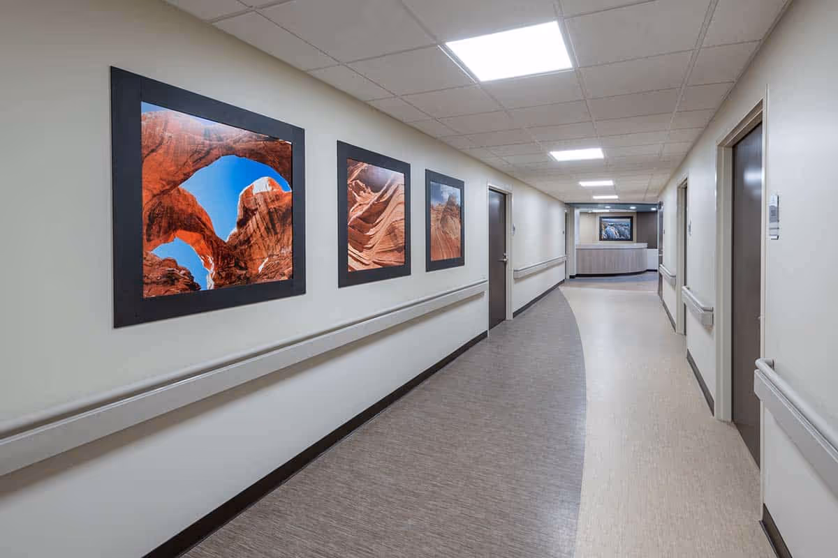 A long, well-lit hallway in a senior living facility with framed landscape photos along one wall and doors to rooms.
