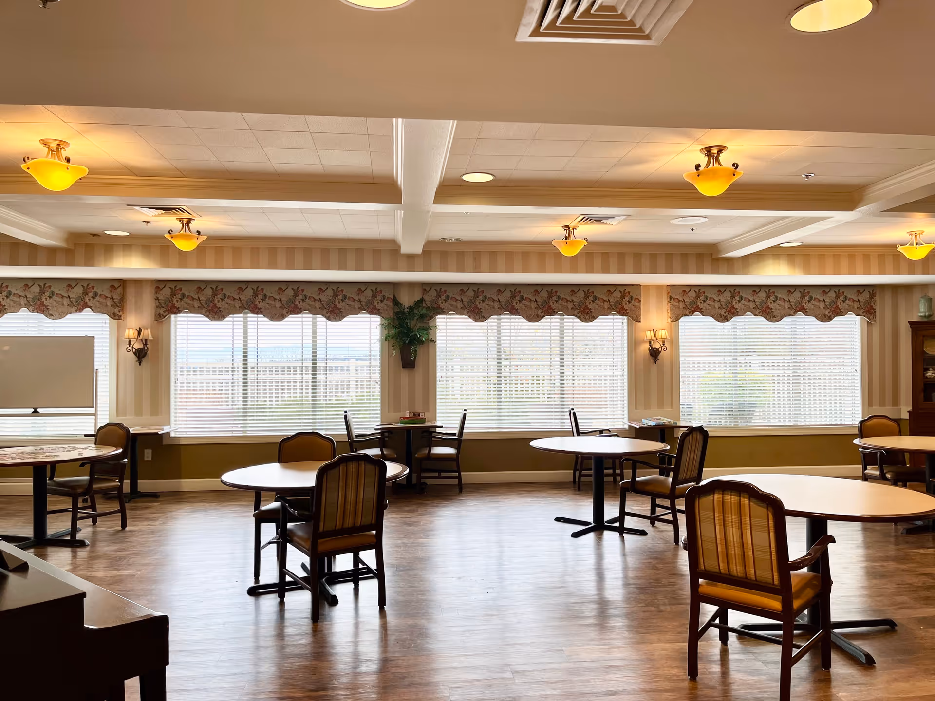 A spacious and well-lit dining room with round tables and cushioned chairs arranged neatly. Large windows with floral valances allow natural light to fill the room. The ceiling features recessed lighting and decorative light fixtures, and the floor is wood. There is a whiteboard and a potted plant near the windows.