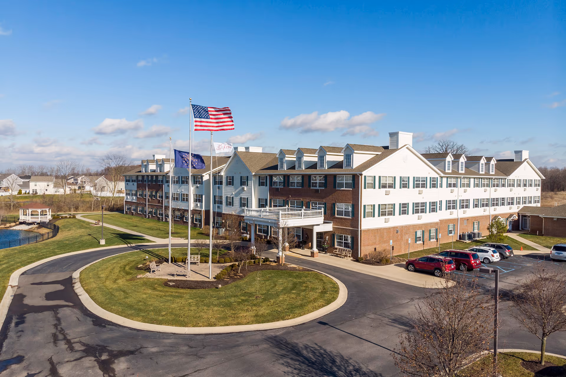 Front exterior of a three-story senior living building with flags, a circular driveway, and parked cars.
