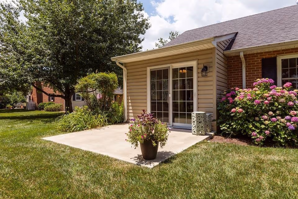 Outdoor patio area with a concrete slab, a potted plant with flowers, and a sliding glass door leading into a building. The building has beige siding and brick walls with a window and flowering bushes nearby. There is a green lawn and trees in the background under a partly cloudy sky.