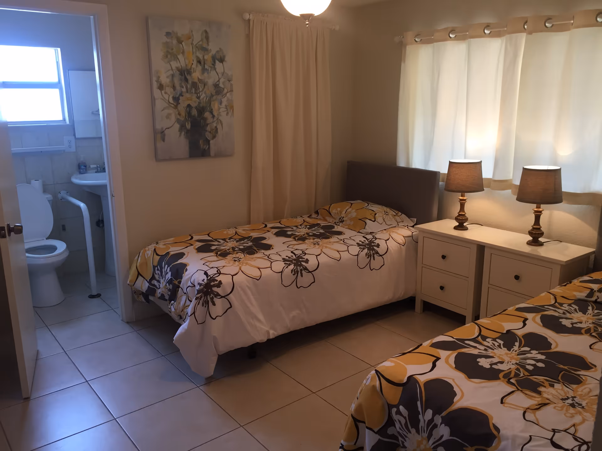 A bedroom with two single beds covered in floral patterned bedspreads in yellow, white, and gray. Between the beds is a white nightstand with two lamps. A window with white curtains is on the right wall. On the left side, there is an open door leading to a small bathroom with a toilet and sink.