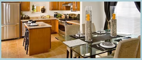 A modern kitchen and dining area featuring wooden cabinets, a stainless steel refrigerator, a kitchen island with bar stools, and a glass dining table set with plates and tall decorative vases filled with sand and candles. The room is well-lit with natural light coming through the window with blinds and curtains.