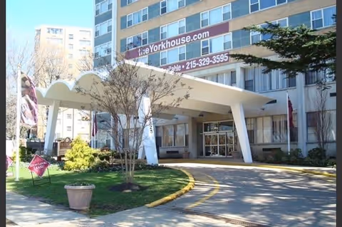 Front exterior view of York House Apartments building with a covered entrance, a curved driveway, some trees, and a sign displaying the website and phone number.