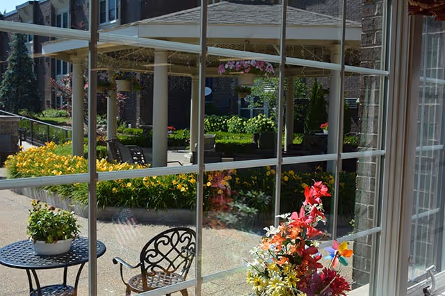 View through a large window showing an outdoor patio area with a round metal table and chairs, potted plants, colorful flowers, and a covered seating area with columns and hanging flower baskets.