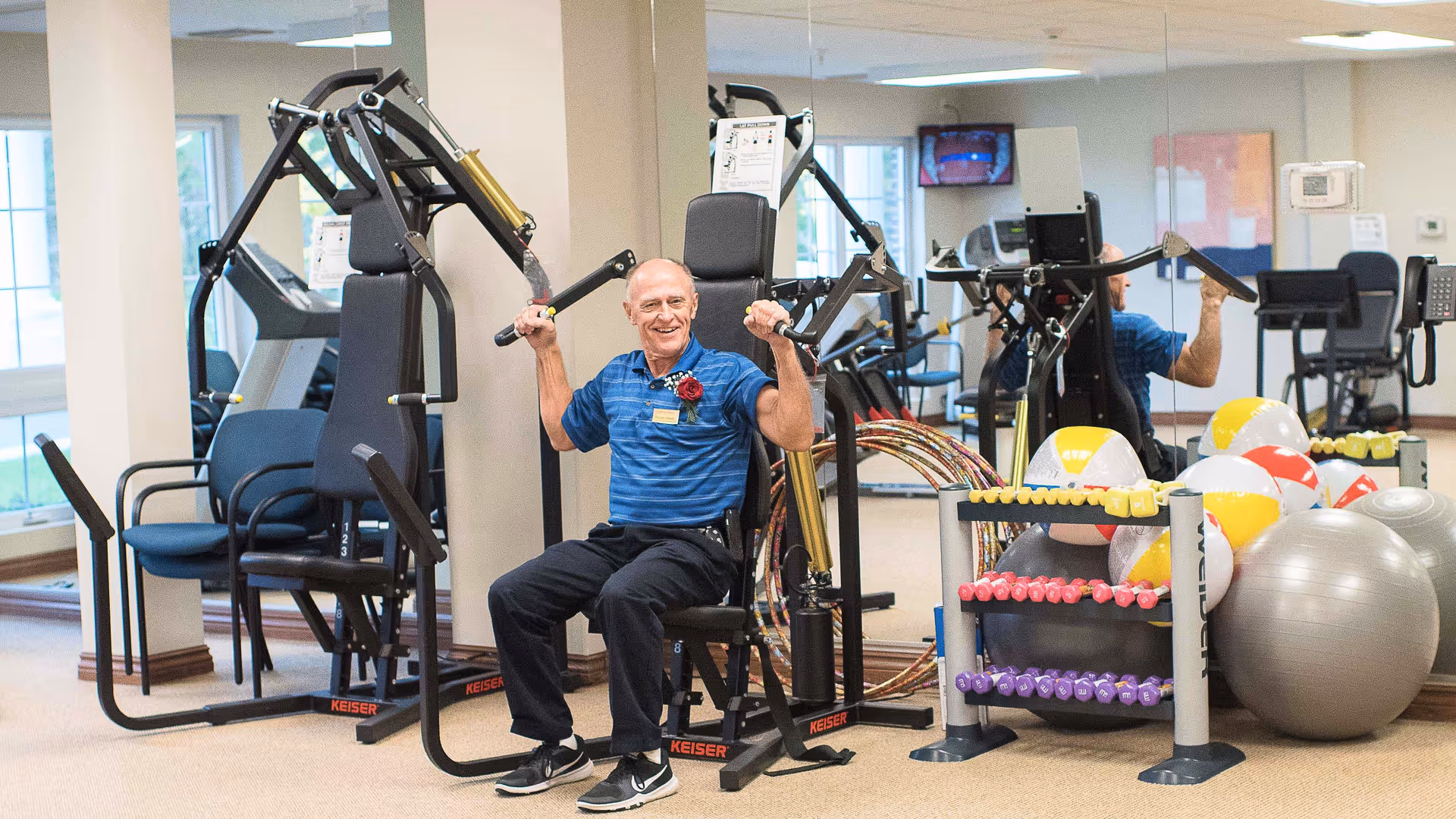 An elderly man in a blue polo shirt and black pants is seated on a strength training machine in a fitness room. He is smiling and holding the machine's handles as if exercising. The room has large windows, mirrors on the wall, various exercise equipment including stability balls, dumbbells, and hula hoops.