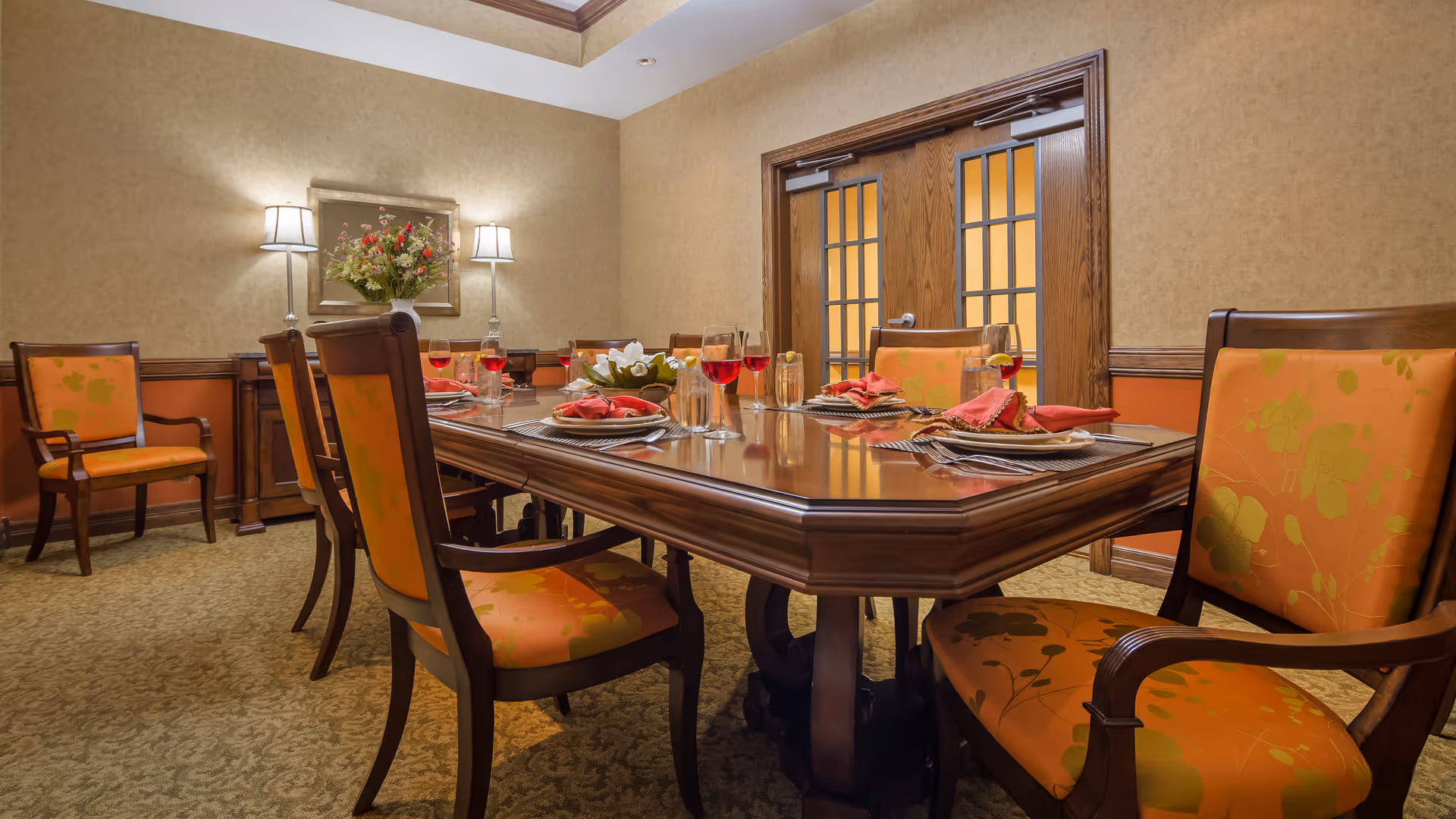 A formal dining room with a large wooden table set for a meal. The table has orange cushioned chairs around it, each place setting includes a plate with a red napkin, a glass of water with lemon, and a glass of red beverage. The room has beige walls with wooden trim, two wall lamps, a floral arrangement on the table, and double wooden doors with glass panels.