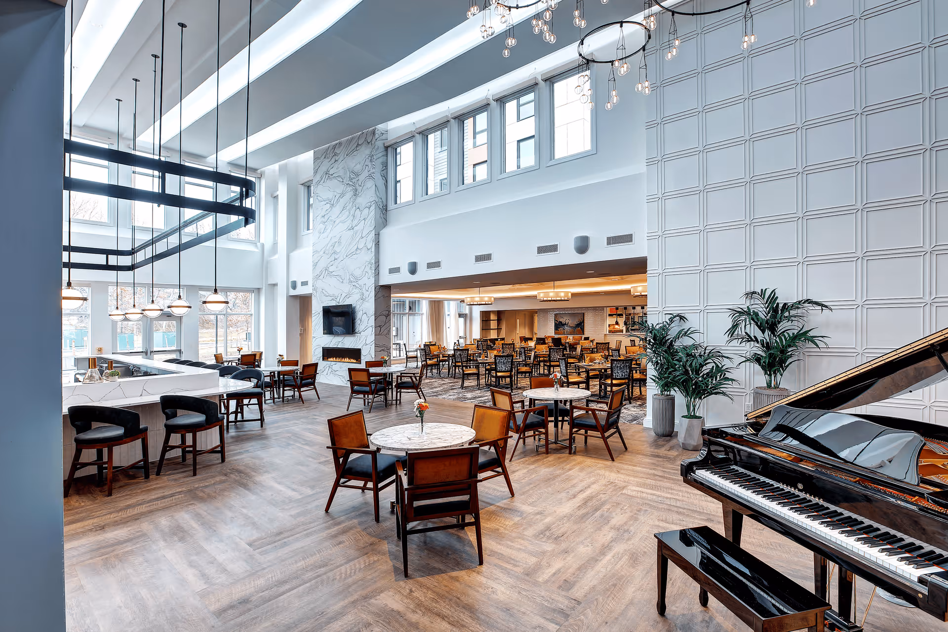 Bright, spacious dining and lounge area with multiple tables and chairs, a grand piano, tall windows, and modern lighting.