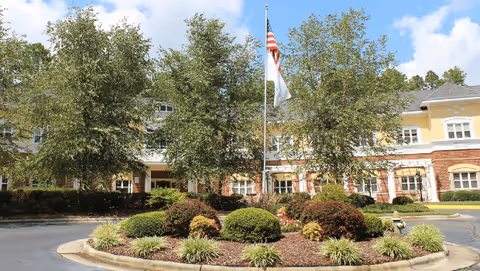 Front exterior view of Fox Hollow Senior Living Community building with a landscaped circular driveway featuring bushes, plants, and two flagpoles with the American flag and another flag, under a partly cloudy sky.