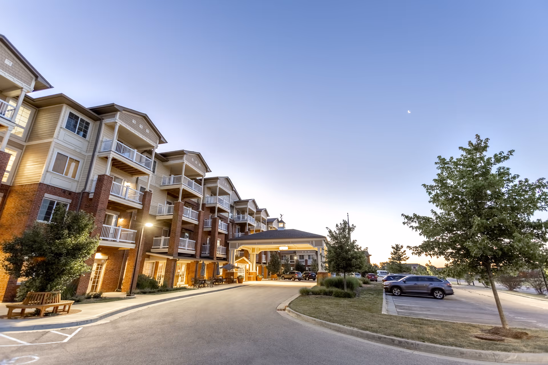 Exterior view of a multi-story senior living facility building at dusk with balconies, a covered entrance, parked cars, trees, and a clear sky with a crescent moon.