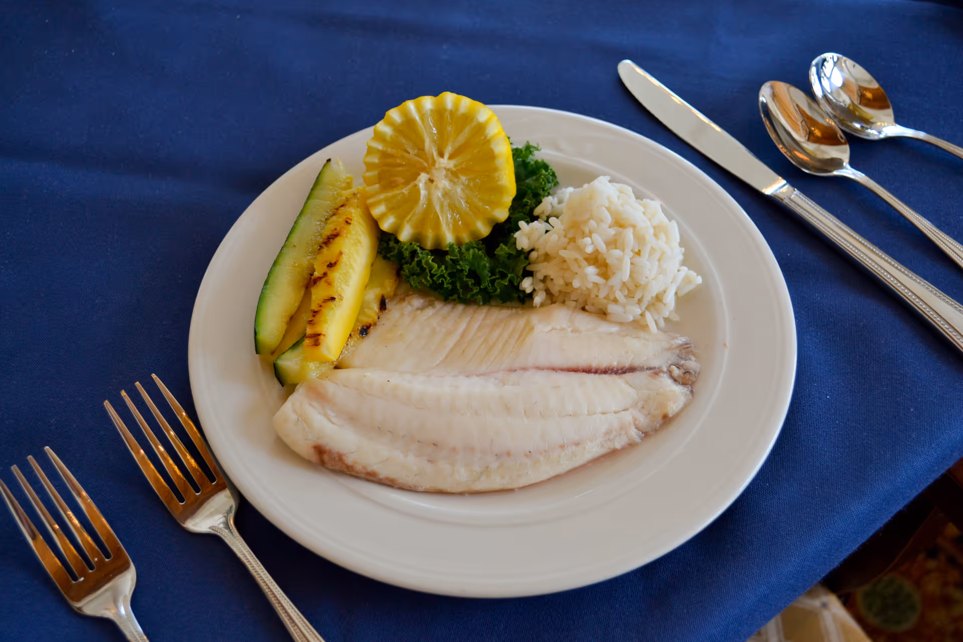 A white plate with a serving of cooked white fish fillet, a portion of white rice, grilled zucchini slices, a lemon slice, and a garnish of leafy greens on a blue tablecloth. Silverware including two forks, a knife, and two spoons are placed around the plate.