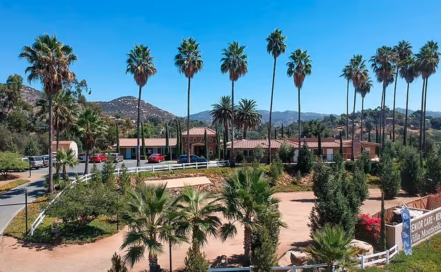 Front view of Villa Monticello senior living facility with a single-story building, tall palm trees, landscaped grounds and mountains in the background.