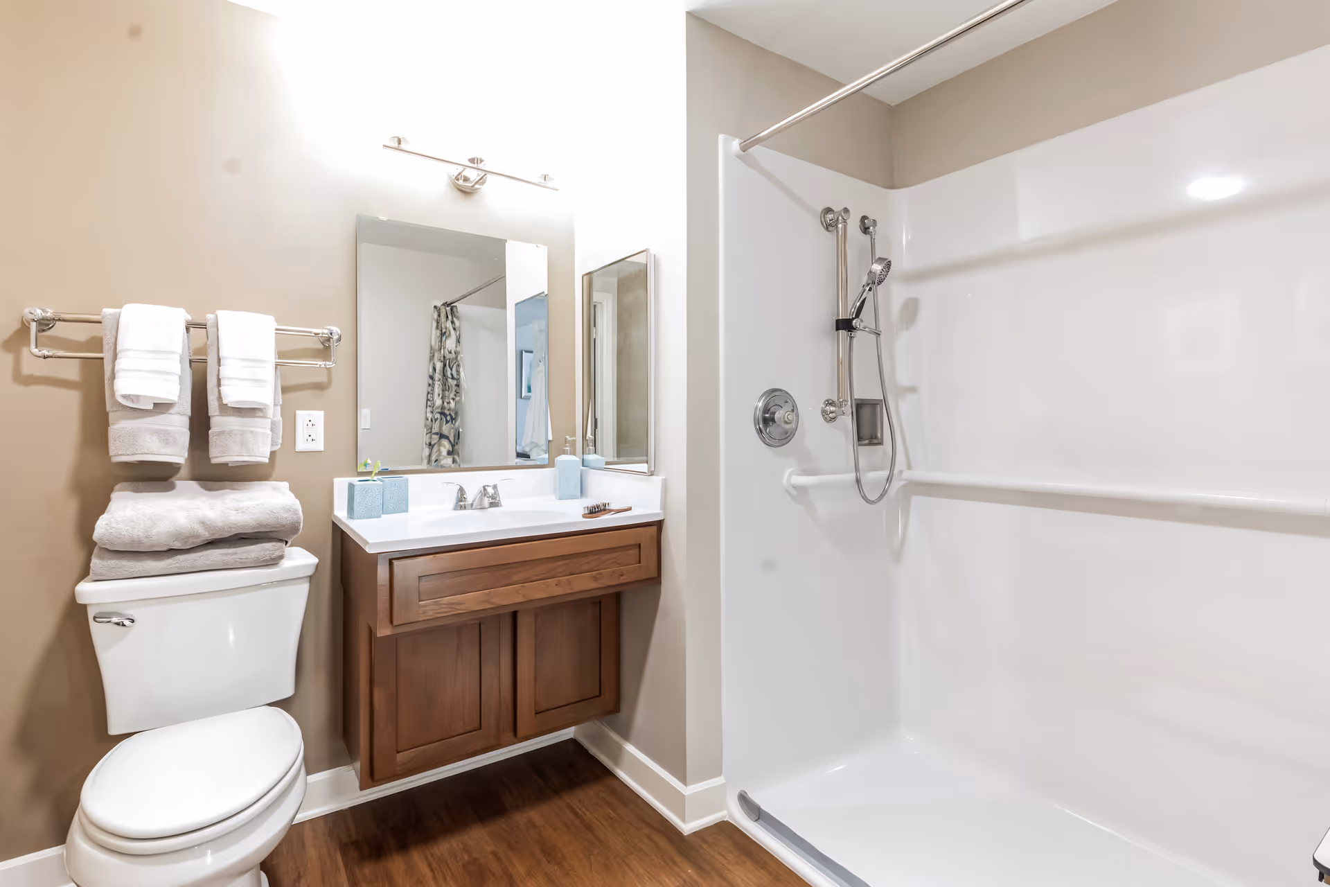 Bright accessible bathroom with a toilet and towel rack beside a vanity and mirror on the left and a walk-in shower with grab bars and handheld showerhead on the right.