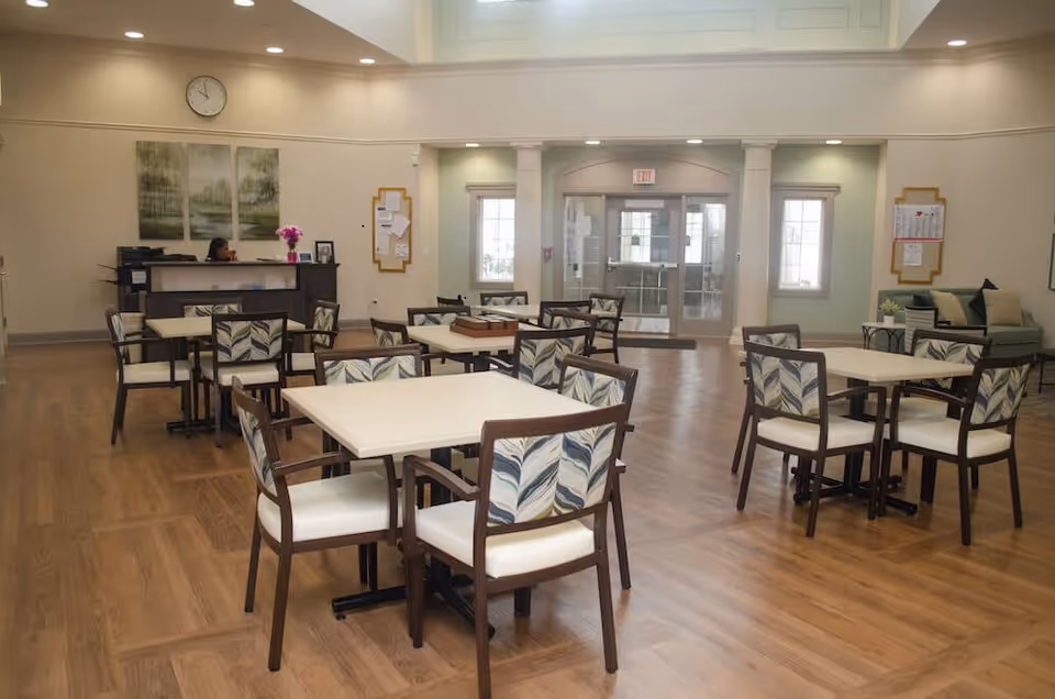 A spacious, well-lit common area with multiple square tables and chairs featuring patterned backs. The room has wooden flooring, a reception desk with a person seated behind it, wall art, a clock, and a seating area with a couch and cushions near the entrance doors.