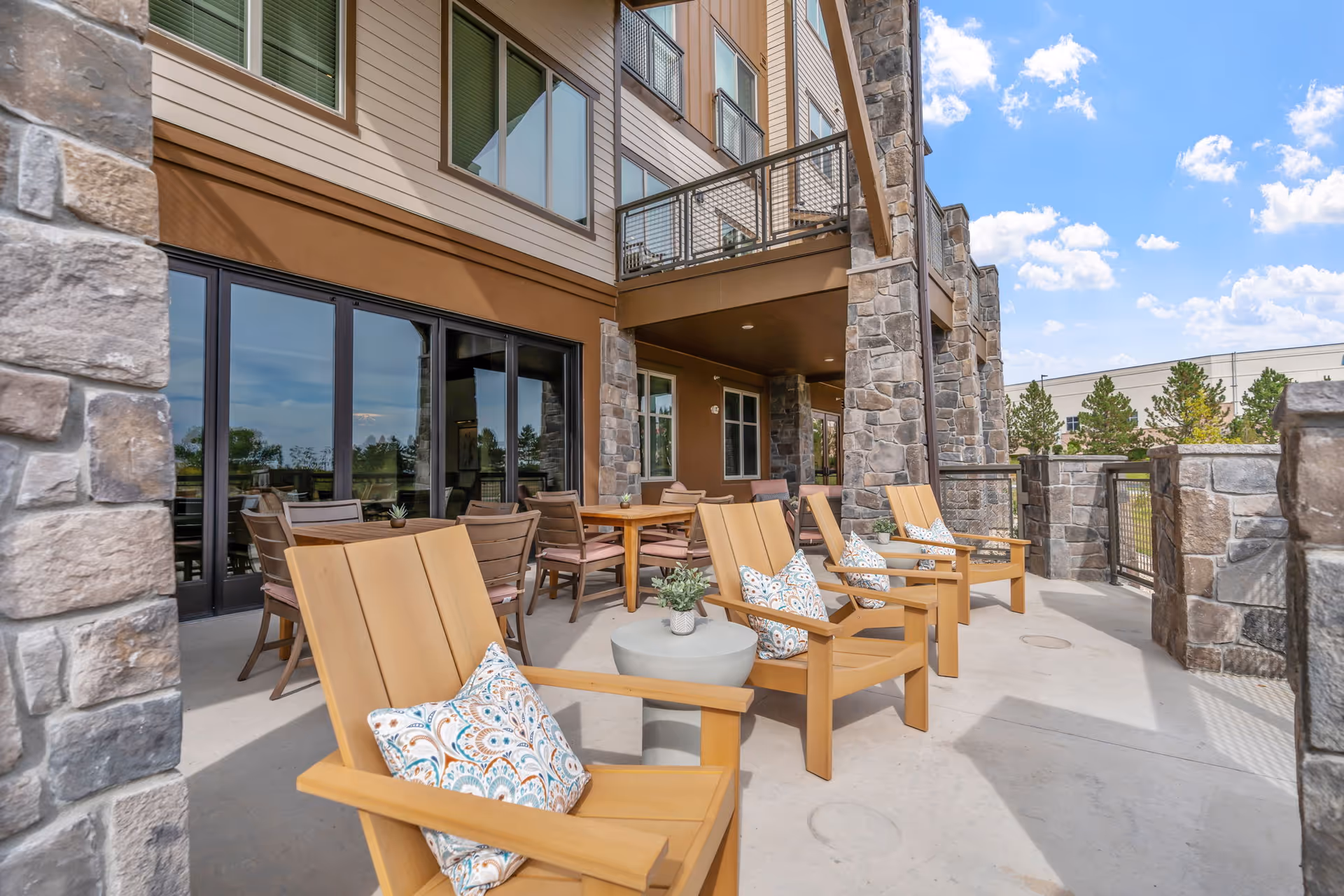 Outdoor patio with wooden lounge chairs and patterned cushions, tables, and a stone-clad building façade under a blue sky.