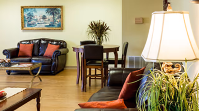 A cozy living room area with two dark leather sofas adorned with red and orange cushions, a wooden coffee table, a small round dining table with four chairs, a large table lamp, a potted plant, and a framed painting on the wall.