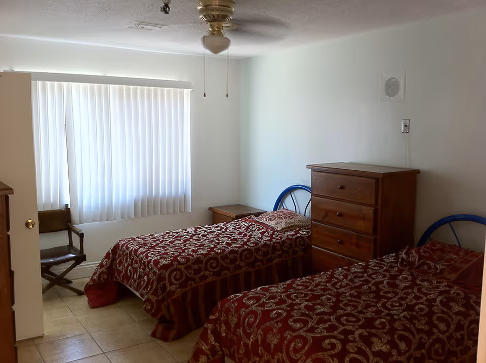 Two twin beds with red patterned bedding, dresser, chair, and window blinds.