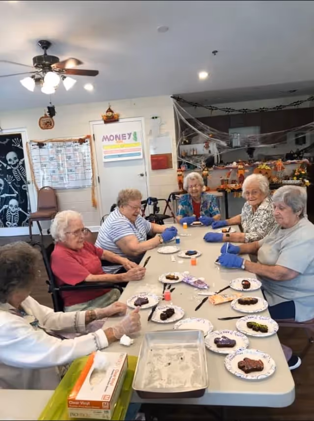 A group of elderly women sitting around a table in a communal room, engaging in a craft or baking activity. They are wearing gloves and appear to be decorating or preparing small cakes or cookies on paper plates. The room is decorated with Halloween-themed items, including a skeleton poster and spider webs. There is a ceiling fan overhead and a kitchen area in the background.