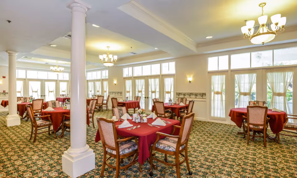 A spacious dining room with multiple round tables covered with red tablecloths, each set with napkins, silverware, and small flower arrangements. The room has large windows with sheer curtains allowing natural light to fill the space, patterned carpet flooring, and elegant chandeliers hanging from the ceiling.