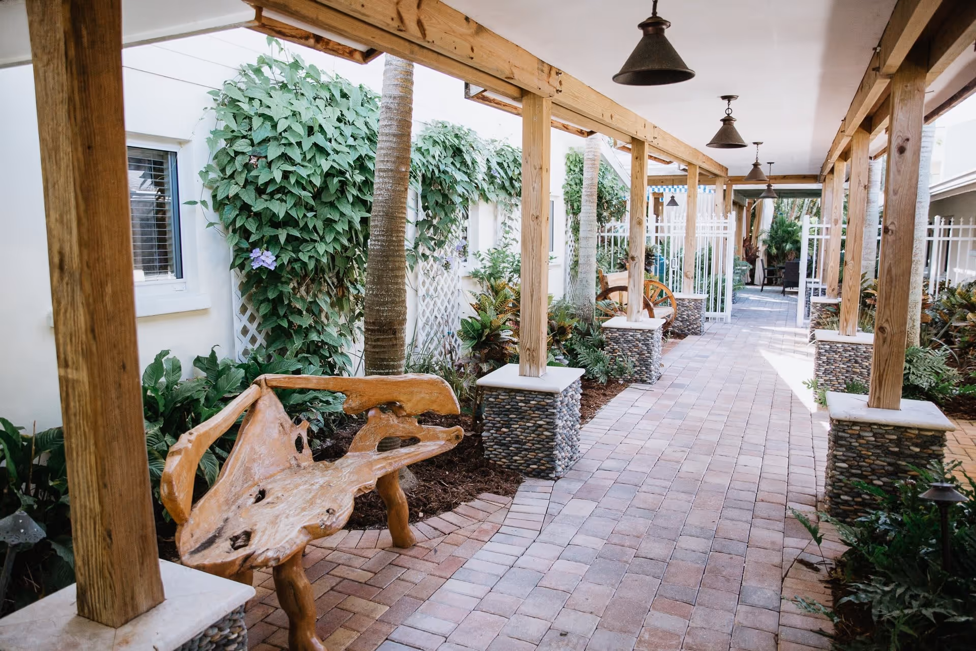 Covered outdoor walkway with stone pillars and wooden beams, featuring a unique wooden bench and lush green plants along the sides, leading to a white gate and seating area in the background.