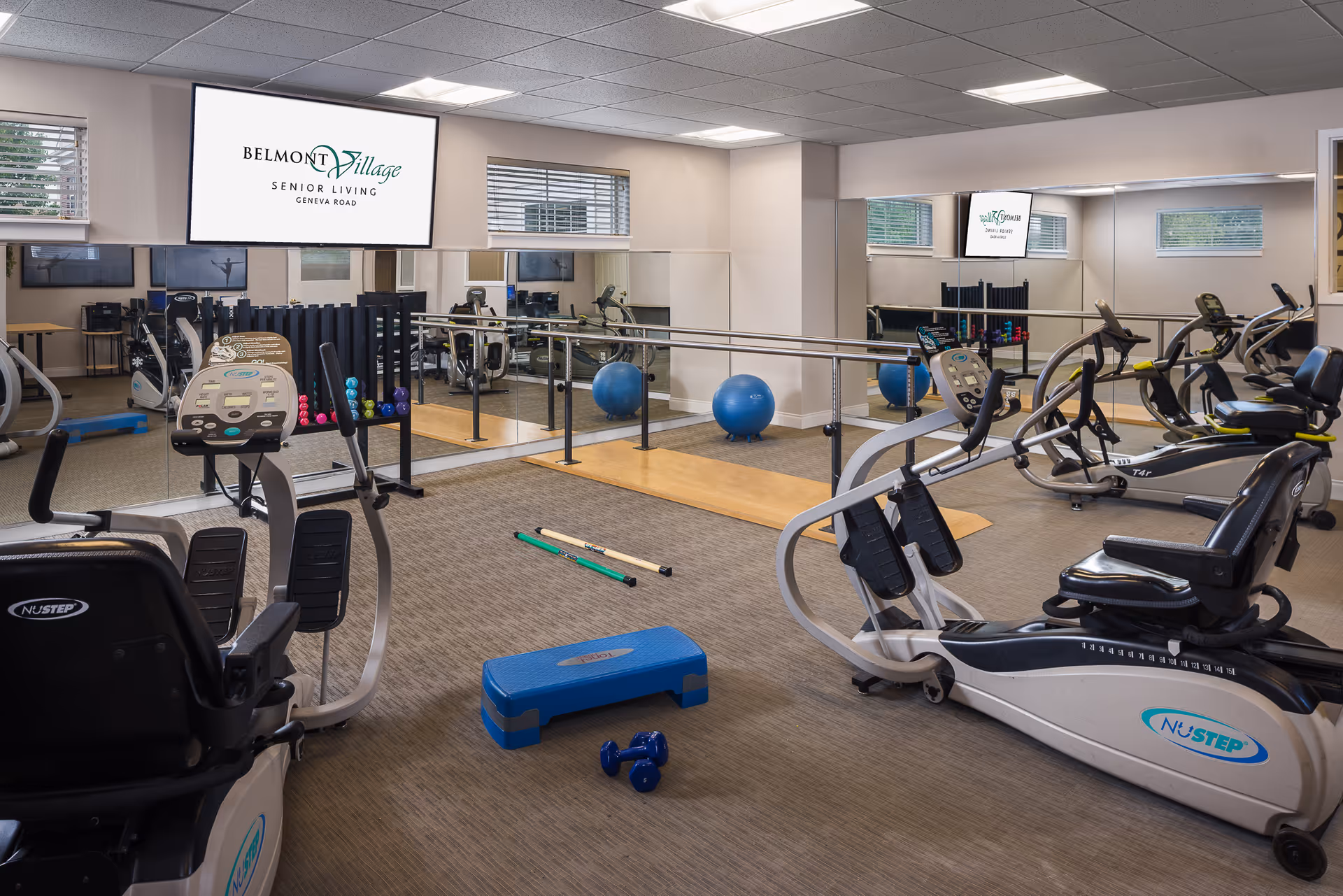 Interior view of a senior living facility exercise room with various fitness equipment including recumbent bikes, dumbbells, a step platform, exercise balls, and hand weights. The room has large mirrors on the walls, windows with blinds, and a television screen displaying the Belmont Village Senior Living Geneva Road logo.