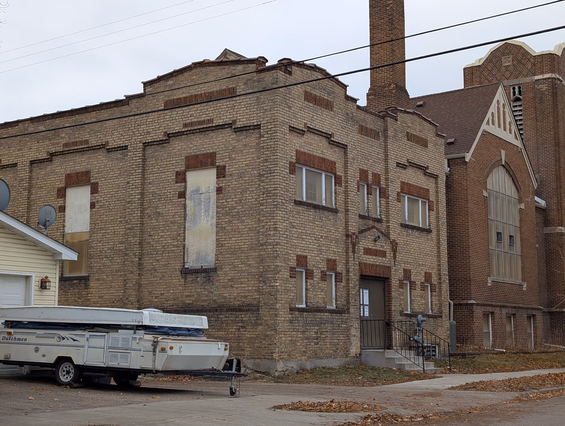 Exterior view of a brick building with boarded-up windows and a closed door with stairs leading up to it. A pop-up camper trailer is parked on the driveway beside the building. The sky is overcast and there are some fallen leaves on the ground.