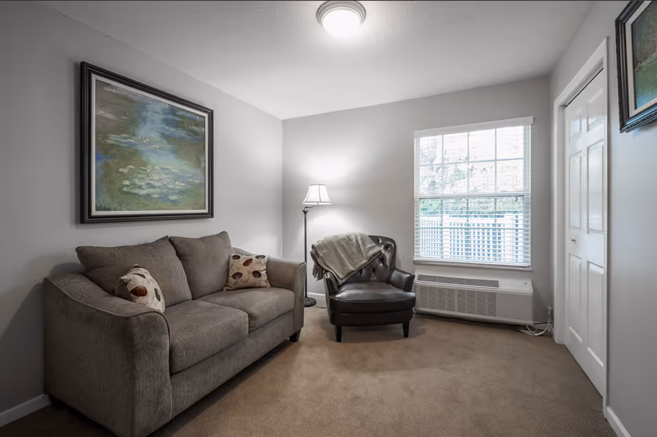 Small living room with a gray sofa, leather armchair, floor lamp, framed artwork, and a window with blinds.