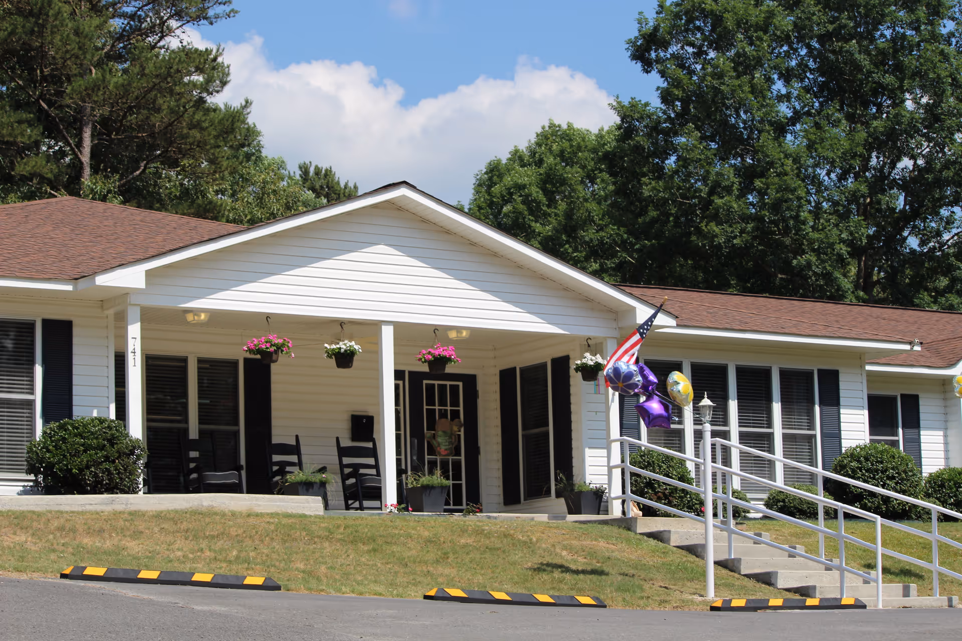 Exterior view of a single-story white building with a brown roof, featuring a covered porch with black rocking chairs and hanging flower pots. There is a ramp with a white railing leading to the entrance, decorated with an American flag and colorful balloons. The building is surrounded by green bushes and trees under a partly cloudy sky.