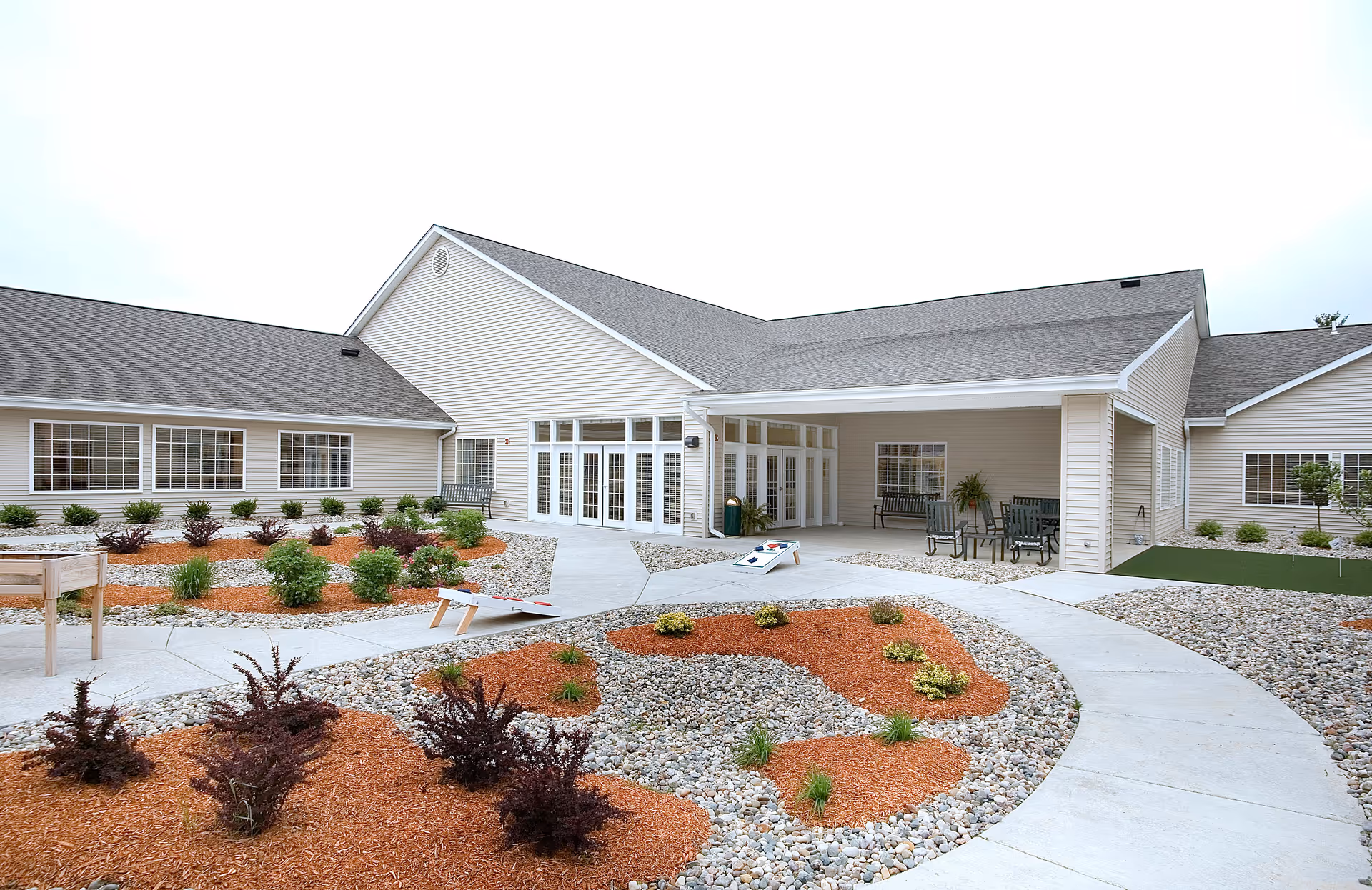 Outdoor courtyard area of The Oaks at NorthPointe Woods featuring a landscaped garden with mulch and rocks, a concrete walkway, two cornhole game boards, and a covered patio with benches and a table with chairs.
