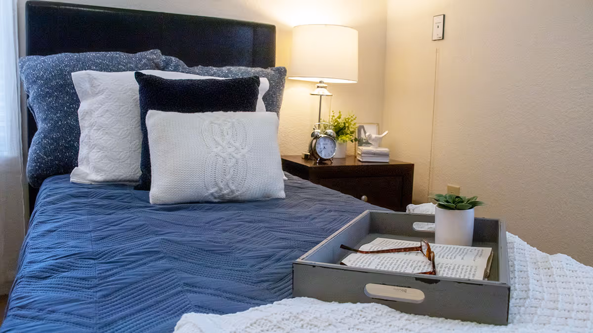 A neatly made bed with a dark blue quilt and multiple pillows in shades of white and dark blue. A gray tray on the bed holds an open book, a pair of glasses, and a small potted plant. Next to the bed is a wooden nightstand with a table lamp, an alarm clock, a small plant, and some decorative items. The room has beige walls and a cozy, inviting atmosphere.