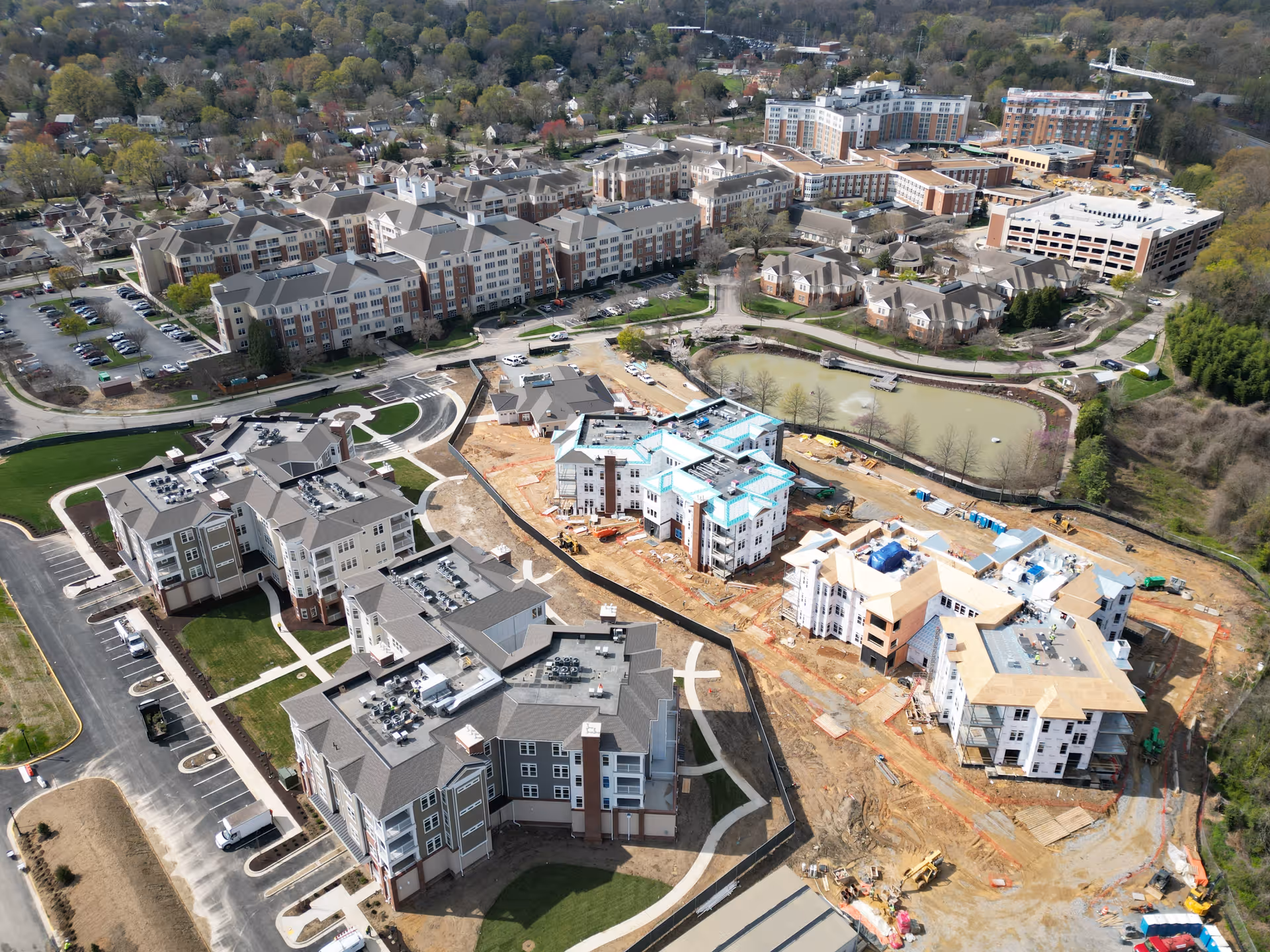 Aerial view of Westminster Canterbury Richmond senior living facility showing multiple large residential buildings, parking lots, a pond, and ongoing construction of new buildings surrounded by trees and a nearby neighborhood.