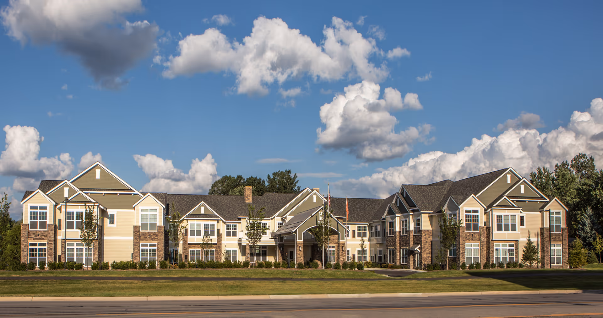 Exterior view of a large two-story senior living facility building with beige and brown siding, multiple windows, and a central entrance under a clear blue sky with scattered clouds.