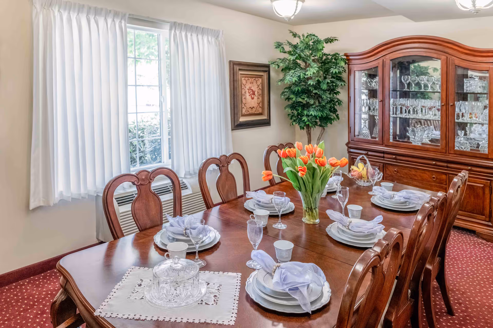 A formal dining room with a wooden dining table set for six people. The table has white plates, cups, glasses, and napkins with napkin rings. A vase with orange tulips is placed in the center of the table. Behind the table is a wooden china cabinet filled with glassware. There is a large window with white curtains and a framed picture on the wall next to a tall green plant.