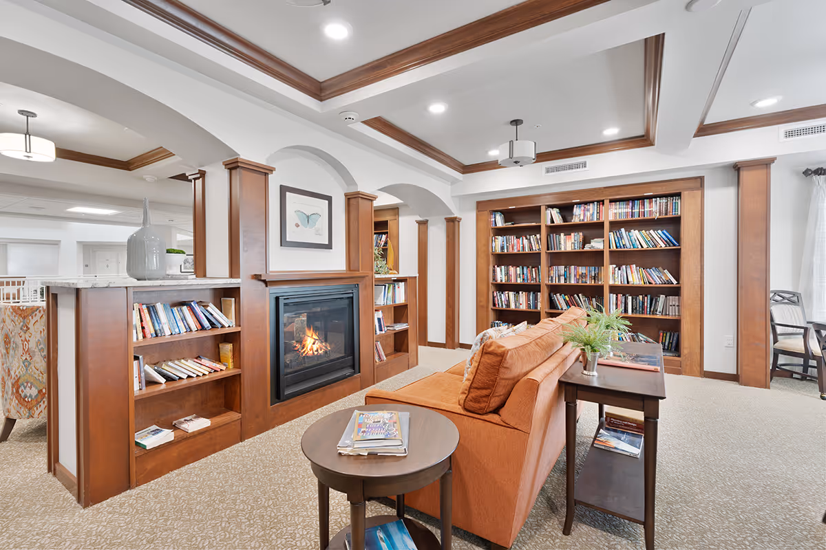 A cozy living room area in a senior living facility featuring a lit fireplace surrounded by built-in wooden bookshelves filled with books. An orange sofa faces the fireplace, with a small wooden side table holding magazines and a plant. The room has a beige carpet, white walls with wooden trim, and recessed lighting in the ceiling. In the background, there is a dining area with chairs and a large vase on the counter.