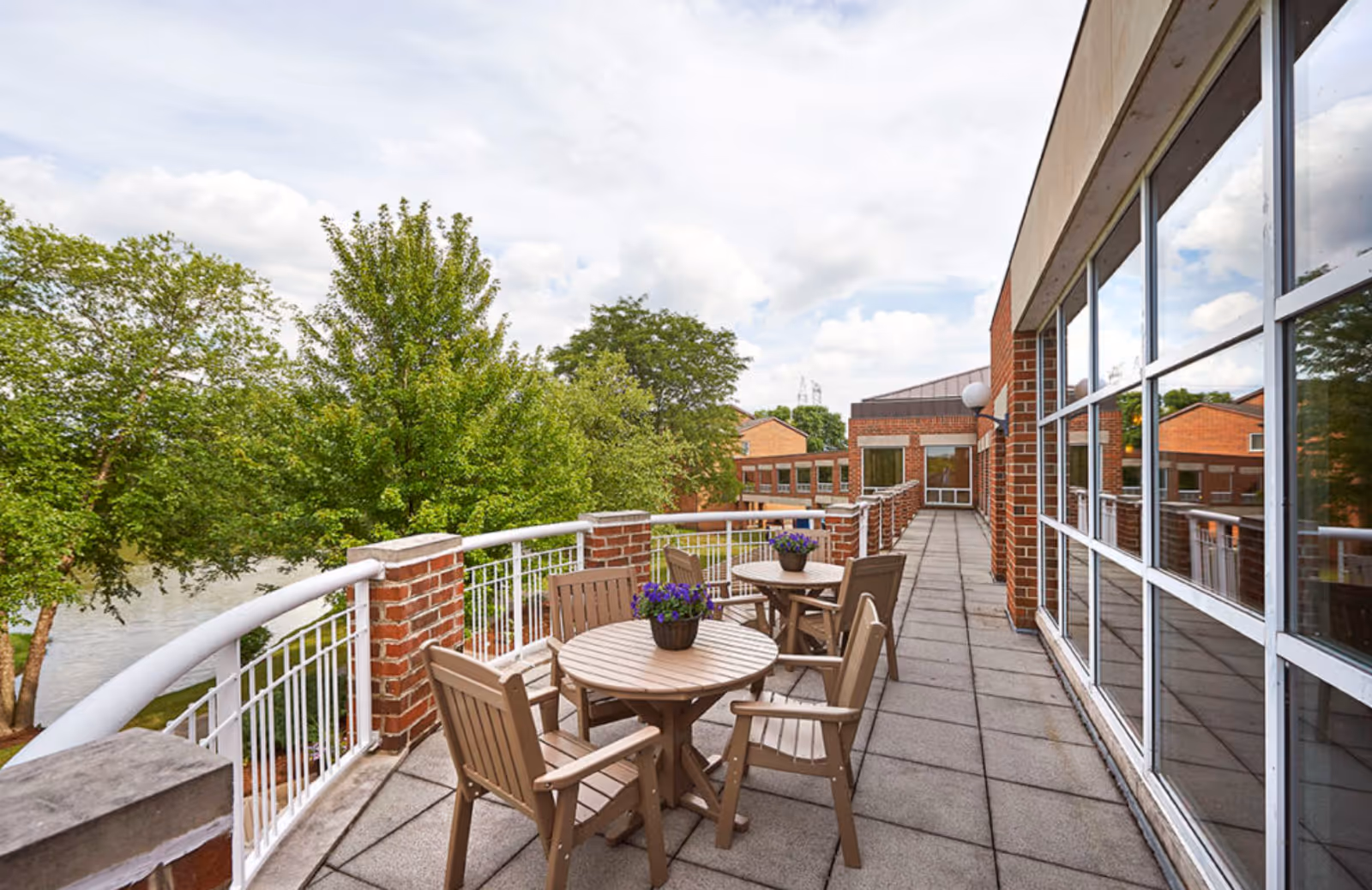 Outdoor terrace with round tables and chairs along a railing beside a brick building overlooking trees and water.