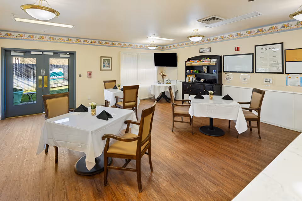 A dining room in a senior living facility with four tables covered in white tablecloths, each set with black folded napkins and small flower arrangements. The room has wooden flooring, beige walls with a floral border near the ceiling, and double glass doors leading outside. There is a black cabinet with shelves holding various items, a wall-mounted TV, and several framed notices and boards on the walls.