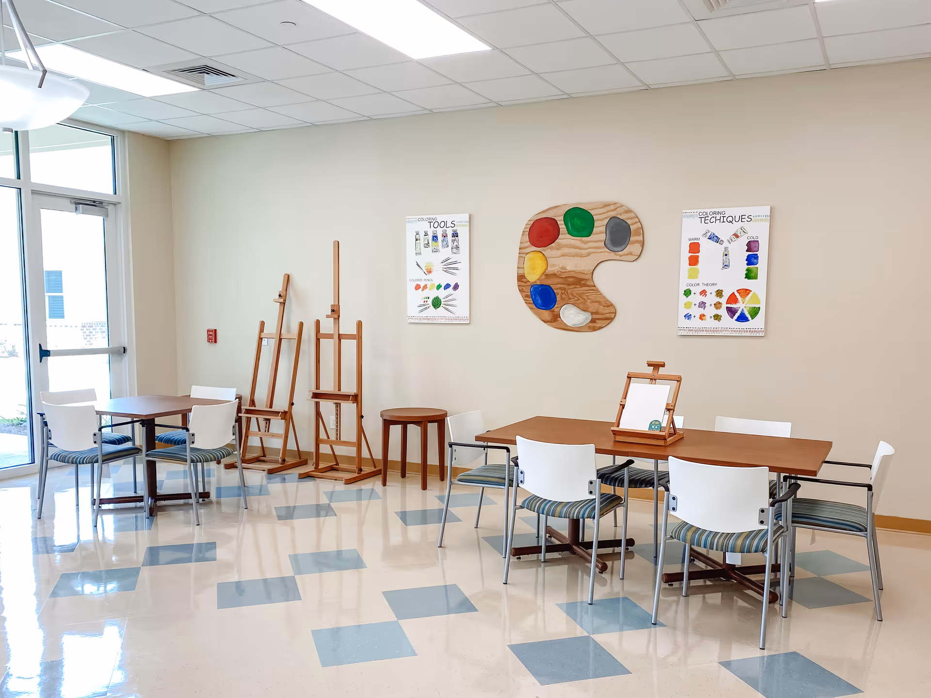Bright activity room with tables and chairs, multiple easels, and a large decorative artist palette on the wall.