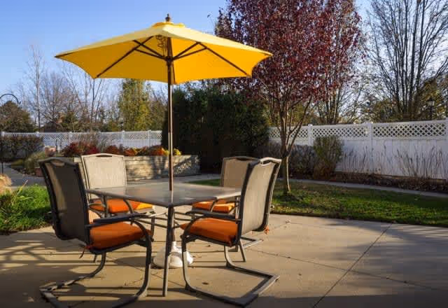 Outdoor patio area with a square table, four chairs with orange cushions, and a large yellow umbrella providing shade. The patio is surrounded by a white fence and landscaped with trees and shrubs.