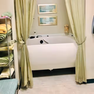 A white walk-in bathtub with a built-in seat and faucet, partially enclosed by light green curtains tied back on either side. To the left, there is a metal shelving unit holding neatly folded towels in various shades of green and yellow. Above the bathtub, two framed pictures with calming words hang on the wall. The floor is carpeted in a light color.