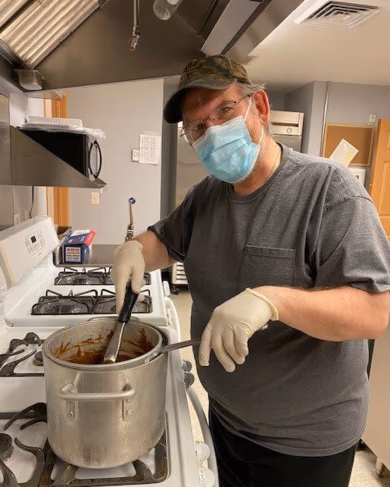 A person wearing a face mask, gloves, and a cap stirs a large pot on a stove in a kitchen.