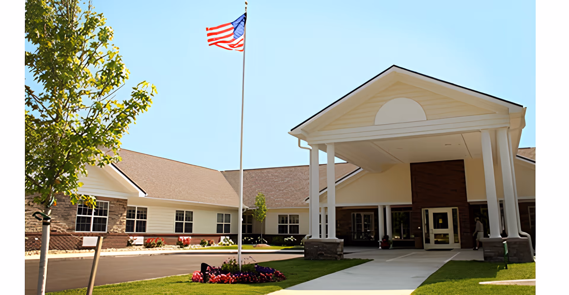 Front entrance of a single-story senior living building with a covered portico, American flag, and landscaped lawn.