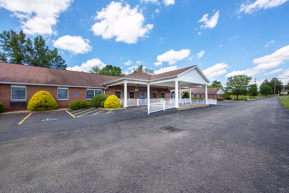 Single-story brick senior living building with a covered front entrance, white railing, and an empty asphalt parking lot under a blue sky.